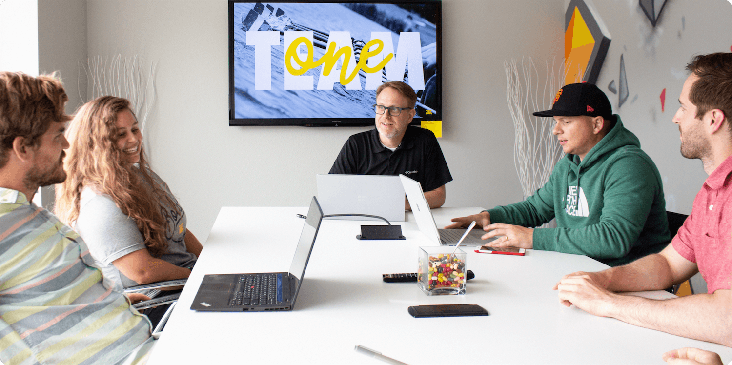 BrainStorm team members gathered around a table in a meeting, with a TV in the background displaying the "One Team" logo.